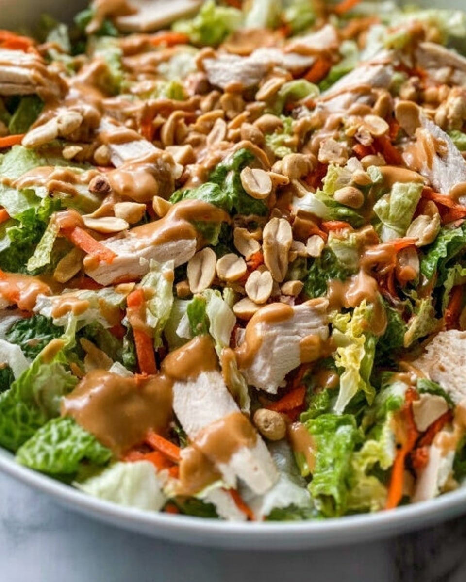 The image shows a close-up of a salad in a white bowl on a white marbled surface. The salad has layers of torn green lettuce leaves, thin orange carrot strips, and pieces of cooked white chicken scattered on top. There are also whole or sliced peanuts sprinkled over the salad. A creamy brown dressing is drizzled evenly across the top, giving a smooth texture contrast to the crisp vegetables and chicken. The mix looks fresh and colorful with green, orange, white, and brown shades all visible. photo taken with an iphone --ar 4:5 --v 7