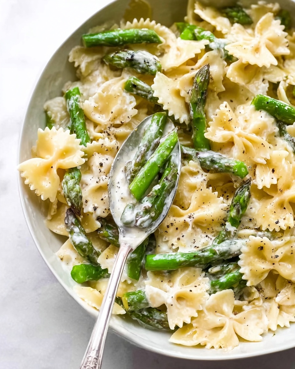 A close-up view of a white plate filled with farfalle pasta mixed with bright green asparagus pieces. The pasta is covered with a creamy white sauce, and some black pepper is sprinkled on top. A silver spoon rests in the middle, holding some pasta and sauce. The plate sits on a white marbled surface. The dish has a simple, fresh look with smooth textures of sauce and firm, crisp asparagus. Photo taken with an iphone --ar 4:5 --v 7