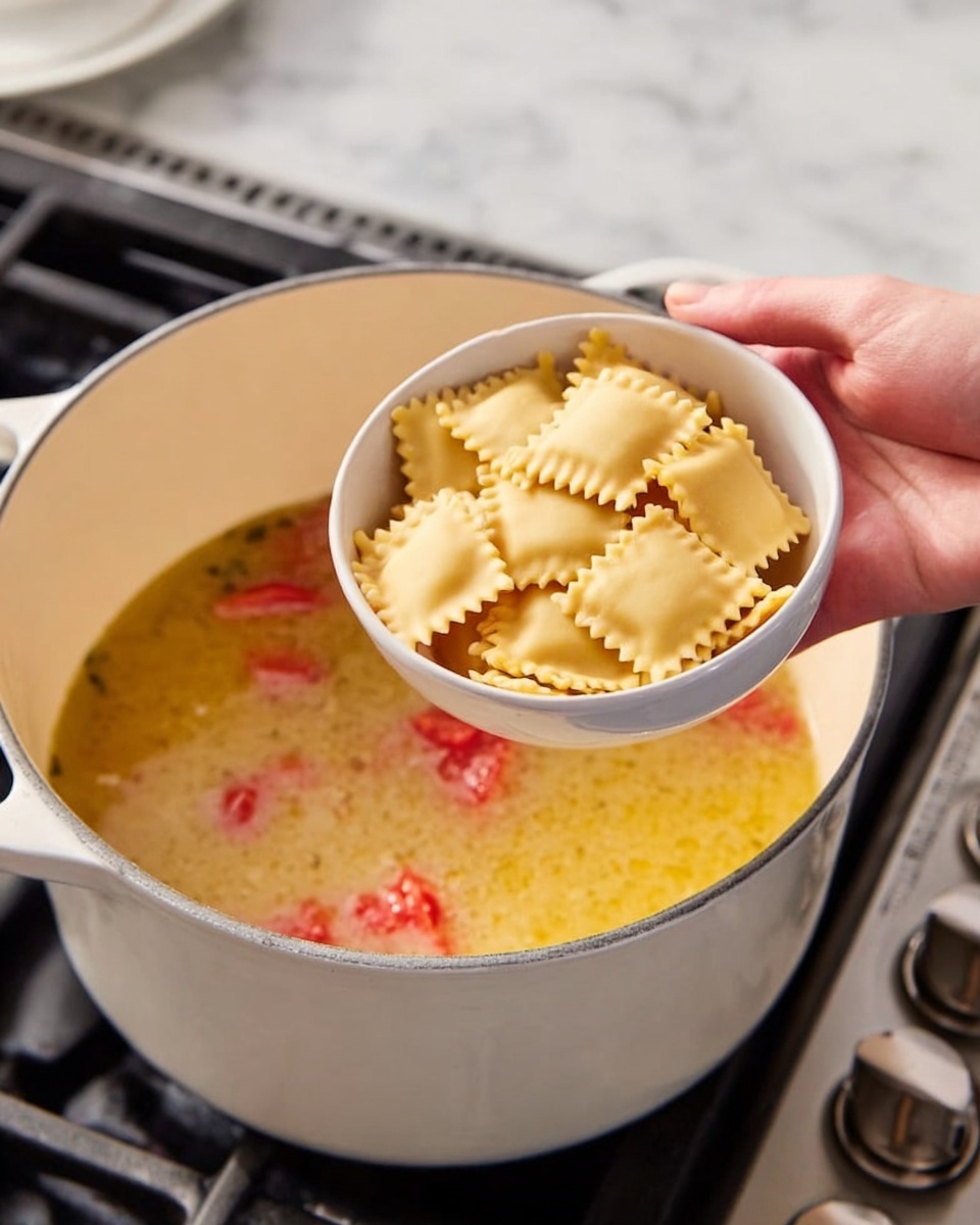 A close-up image shows a woman's hand holding a small white bowl filled with square-shaped ravioli pasta above a large white pot on a stove. The pot contains a light yellow broth with small pieces of red tomatoes floating on top. The pasta is fresh with a smooth surface and fluted edges. The background surface is white marble with some stove knobs visible on the right side. The scene captures the moment just before the ravioli is dropped into the broth. photo taken with an iphone --ar 4:5 --v 7
