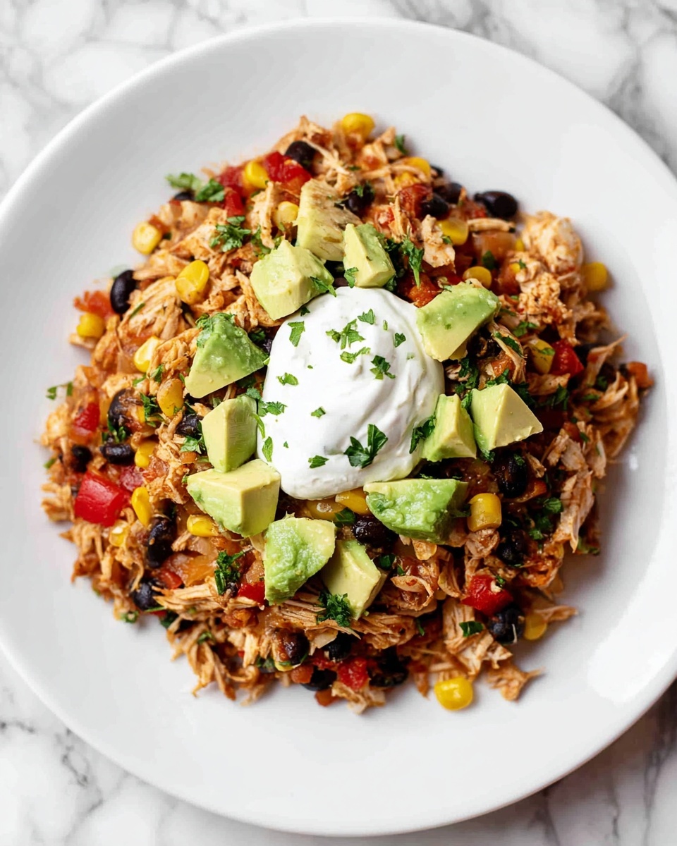 A white plate holds a colorful layered dish with four main layers. The base layer is shredded cooked chicken mixed with finely chopped red peppers, black beans, corn kernels, and small bits of green herbs, creating a textured and varied color mix of light brown, red, black, yellow, and green. On top of this mix, there are scattered chunky pieces of fresh green avocado adding a smooth, soft contrast. The dish is finished with a generous dollop of creamy white sour cream placed in the center. The plate sits on a white marbled surface. Photo taken with an iphone --ar 4:5 --v 7