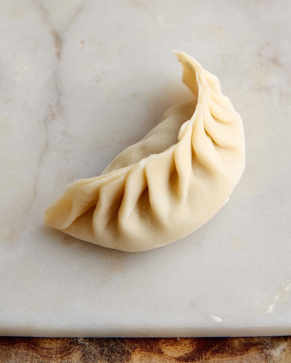 A close-up image of a single folded dumpling on a white marbled surface with a wooden edge at the bottom. The dumpling has a smooth, light beige dough with crimped edges that create a rippled pattern along the curved side, showing careful pleats layered on top of each other. The shape is semi-circular, with the filling fully enclosed inside the soft dough. Photo taken with an iphone --ar 4:5 --v 7