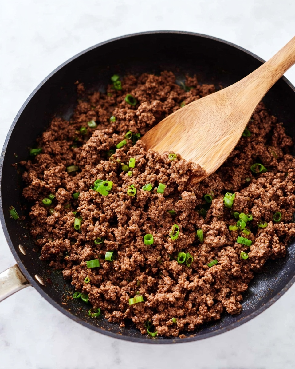 The image shows a black frying pan filled with cooked ground beef that looks brown and crumbly, mixed with small chopped green onions scattered on top for color contrast. A wooden spoon rests inside the pan, partially lifting the beef, showing the texture clearly. The surface under the pan has a white marbled texture, providing a clean and bright background. photo taken with an iphone --ar 4:5 --v 7