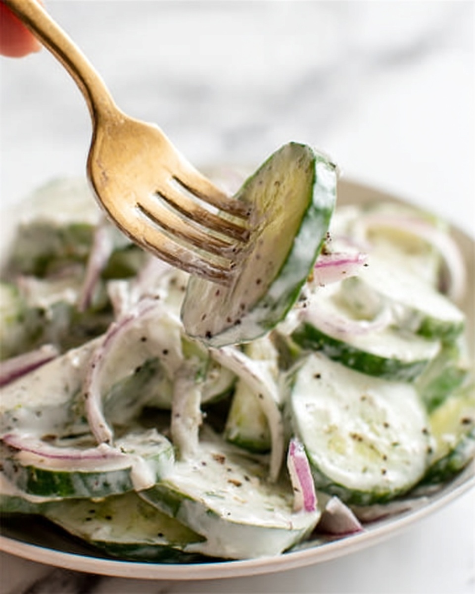 The image shows a close-up of a white plate filled with thick pieces of green cucumbers and light purple slices of onion, both covered in a creamy white dressing with visible bits of black pepper and herbs. A woman's hand holds a gold fork that is piercing a slice of cucumber, lifting it slightly. The background is a white marbled surface, adding a clean and bright feel to the scene. The creamy sauce looks smooth and thick, evenly coating the vegetables, while the onions add a slight shine. photo taken with an iphone --ar 4:5 --v 7