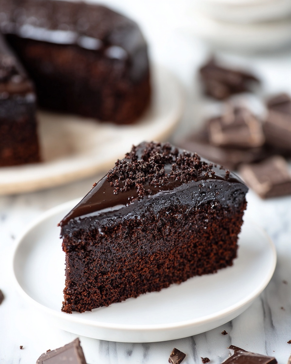 A single slice of rich dark chocolate cake sits on a small white plate, showing a thick, moist, and dense chocolate base layer topped with a shiny, smooth layer of dark chocolate ganache, sprinkled lightly with crumbly chocolate bits on top; in the background, blurred pieces of the whole cake and broken chocolate chunks rest on a white marbled surface, creating a soft and clean look. photo taken with an iphone --ar 4:5 --v 7