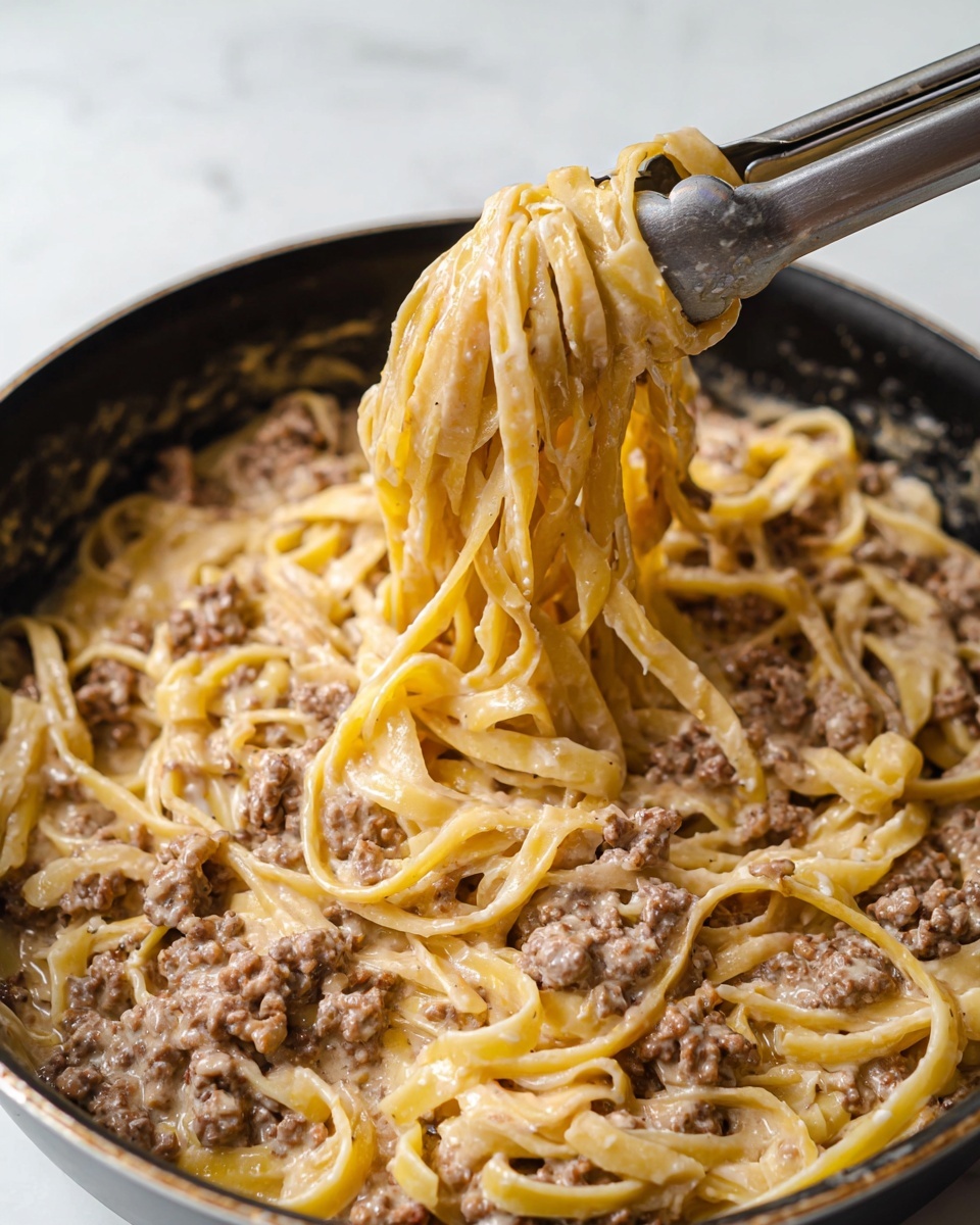 A close-up image shows a large black pan filled with creamy pasta mixed with ground meat. The pasta is long, flat, and yellow, coated in a thick light beige sauce with small brown meat bits spread throughout. Silver tongs hold up a twisted bundle of the pasta and meat from the center of the pan, showing smooth and glossy textures. The background is a white marbled surface. photo taken with an iphone --ar 4:5 --v 7