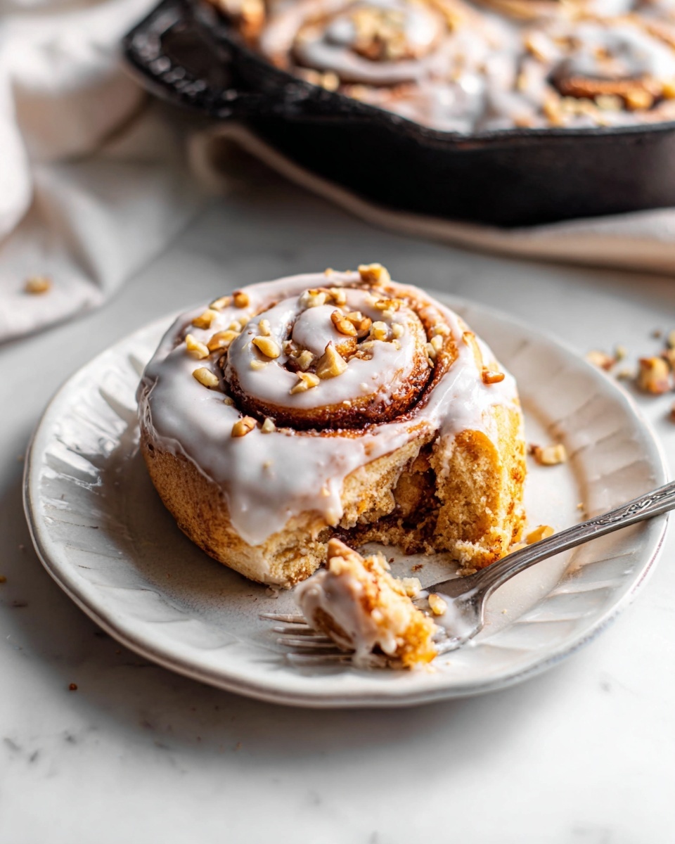A single cinnamon roll rests on a white plate with a slightly ruffled edge, showing its spiral shape made of soft light brown dough layered with cinnamon sugar. The roll is generously topped with a thick, white icing glaze that drips down its sides and is sprinkled with small pieces of chopped nuts, likely walnuts, that add texture and contrast. Part of the roll has a bite taken out, revealing the fluffy and moist inside with a swirl of cinnamon sugar. A silver fork lies on the plate beside the cinnamon roll, and in the background, a black cast iron skillet filled with more cinnamon rolls covered in the same white icing is out of focus. The scene sits on a white marbled surface with soft lighting. photo taken with an iphone --ar 4:5 --v 7