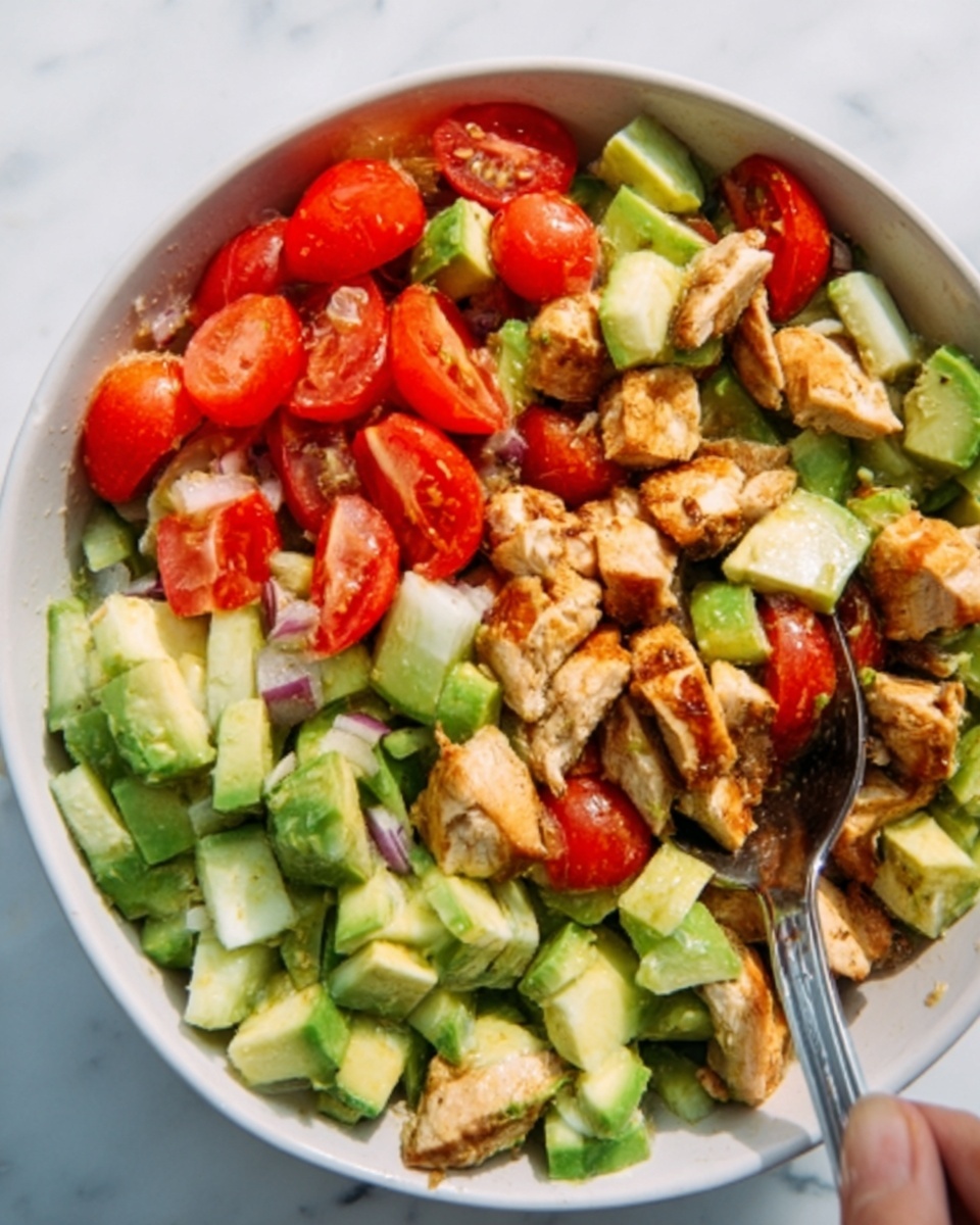 The image shows a colorful salad in a white bowl on a white marbled surface. The salad has several layers: a base of chopped green cucumber pieces with a smooth texture, mixed with bright red cherry tomato halves scattered evenly throughout. On top, there are chunks of light golden-brown grilled chicken with a slightly crispy texture. A woman's hand is holding a spoon on the right side, ready to mix the salad. Photo taken with an iphone --ar 4:5 --v 7