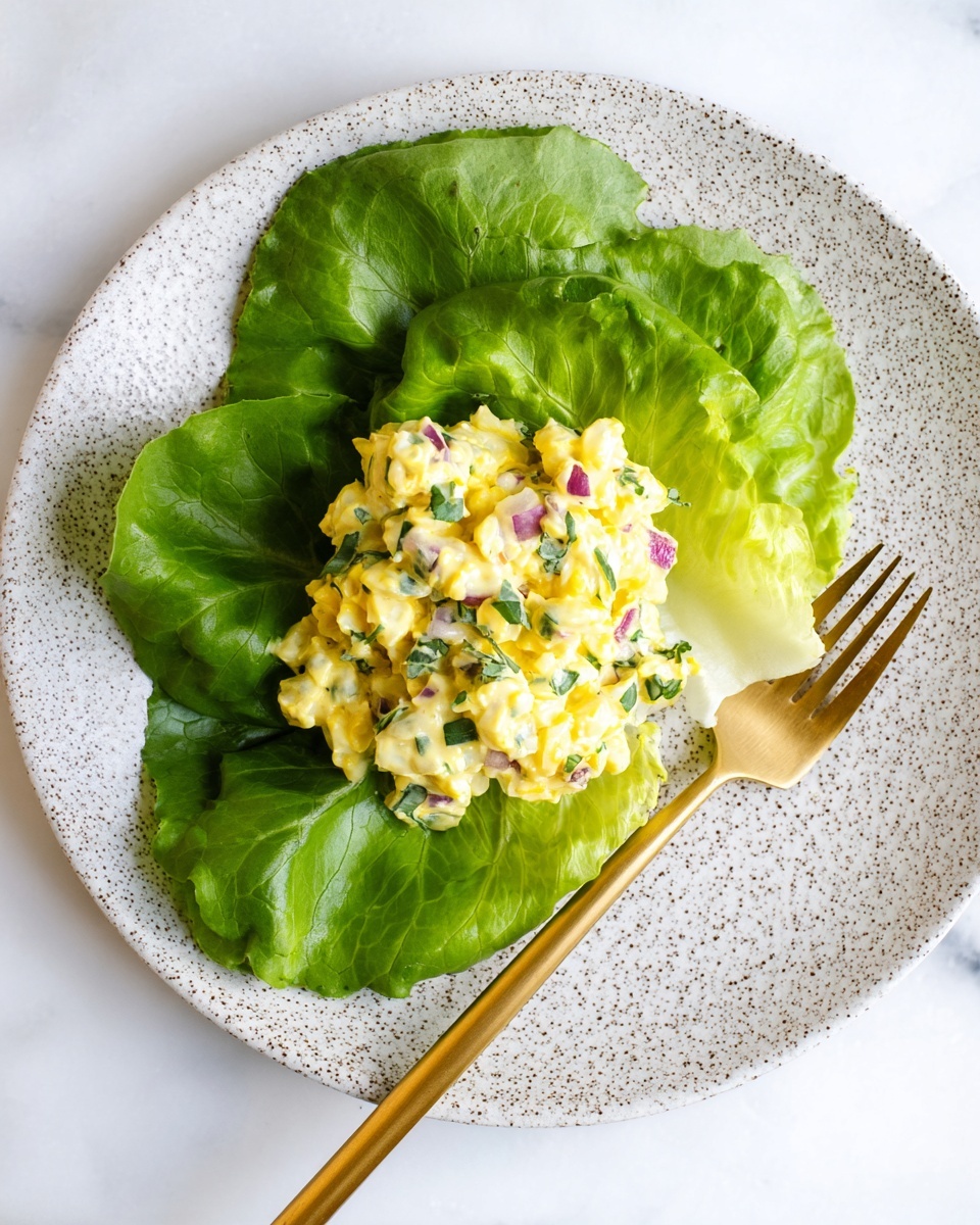 A single white speckled plate sits on a white marbled surface with a gold fork resting on the right side of the plate. On the plate, there are several bright green lettuce leaves arranged in a circular bed. On top of the lettuce, there is a creamy yellow salad made of small egg pieces mixed with finely chopped green herbs and small bits of red onion scattered evenly throughout. The yellow salad forms one thick layer centered on the lettuce leaves, creating a fresh and textured look. photo taken with an iphone --ar 4:5 --v 7