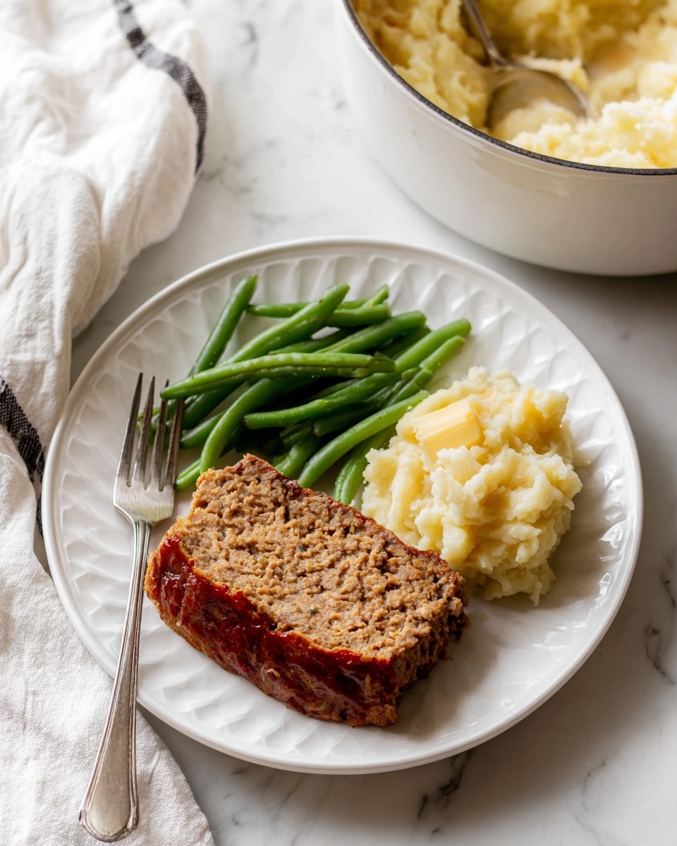 A white plate with three food parts: on the left, a pile of green beans with smooth, shiny green skin; in the middle, a thick slice of brown meatloaf with a slightly crispy, reddish-brown top layer and a dense, crumbly interior; on the right, a scoop of light yellow mashed potatoes with a rough, soft texture. A silver fork rests on the left side of the plate on a white marbled surface. In the background, there is a white pot filled with mashed potatoes, and a white towel with a dark stripe is partly visible near the bottom left. photo taken with an iphone --ar 4:5 --v 7