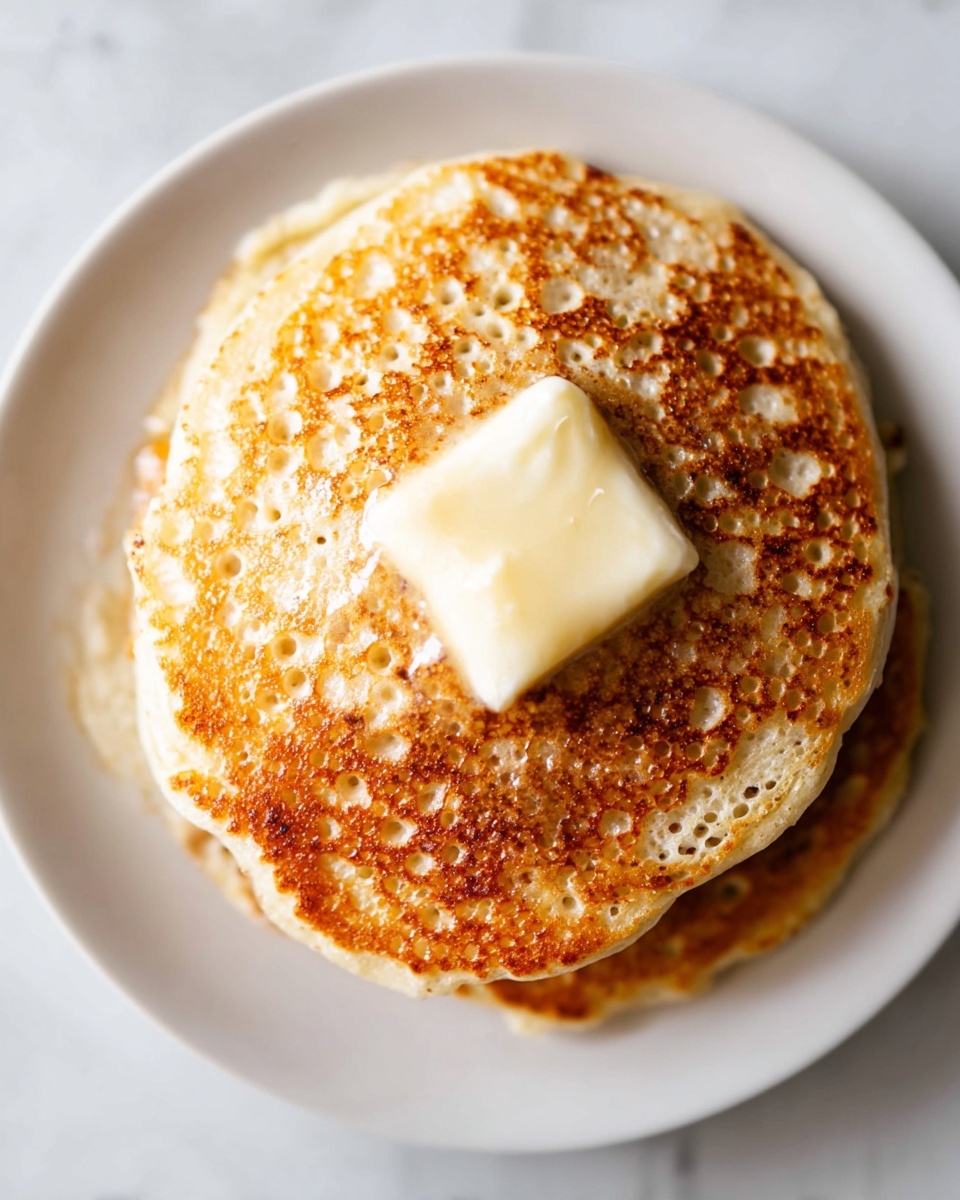 A stack of six thick pancakes sits in the center of a white plate on a white marbled surface. The pancakes are light golden brown with a slightly rough texture, showing a soft inside as a slice is missing from the stack's front. Golden syrup glistens between and around the pancakes, pooling at the base. On top and around the stack, several smooth, pale yellow banana slices add color and freshness. In the background, a white ceramic mug and a small matching white bowl with more banana slices appear softly blurred. Photo taken with an iphone --ar 4:5 --v 7