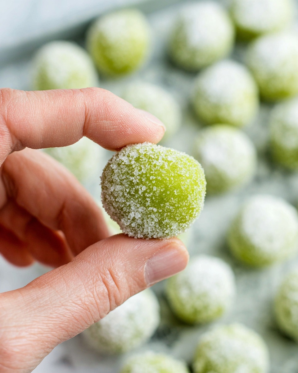 A close-up image shows a woman's hand holding a small round green ball covered mostly in white sugar crystals. The green ball appears smooth underneath the sugar. In the background, more of these green balls coated with sugar are placed close together on a white marbled surface, creating a soft blurry effect. The contrast between the bright green and the white sugar is clear, and the texture of the sugar crystals is rough and sparkly. photo taken with an iphone --ar 4:5 --v 7