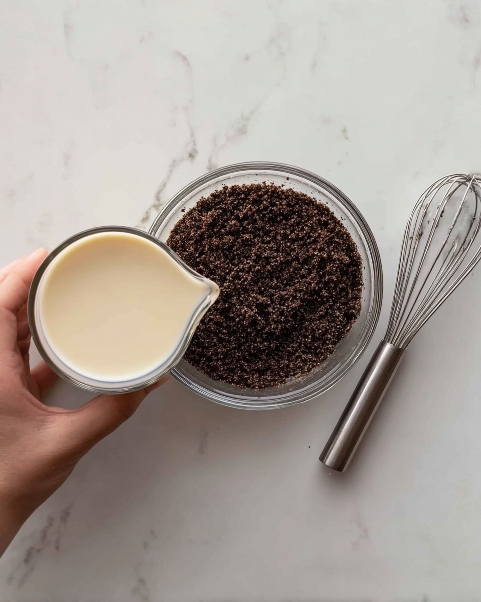A woman's hand holding a small glass jug filled with pale cream-colored liquid over a clear glass bowl containing a dark brown crumbly mixture on a white marbled surface. Next to the bowl on the right side is a metal whisk with a shiny silver handle and thin wires. The dark brown crumbs in the bowl have a rough texture and cover the bottom evenly, creating a neat layer beneath the cream liquid. photo taken with an iphone --ar 4:5 --v 7