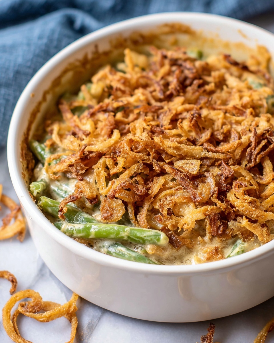 A close-up of a white round bowl filled with creamy baked green bean casserole, showing two main layers: the bottom layer is green beans mixed with creamy sauce in light green and beige tones, and the top layer is a thick, golden-brown crispy fried onion topping that covers the casserole completely. The bowl sits on a white marbled surface with a few crispy onions scattered near it, and a blue cloth is softly blurred in the background. The edges of the bowl have a slight sauce stain, adding a rustic look. Photo taken with an iphone --ar 4:5 --v 7