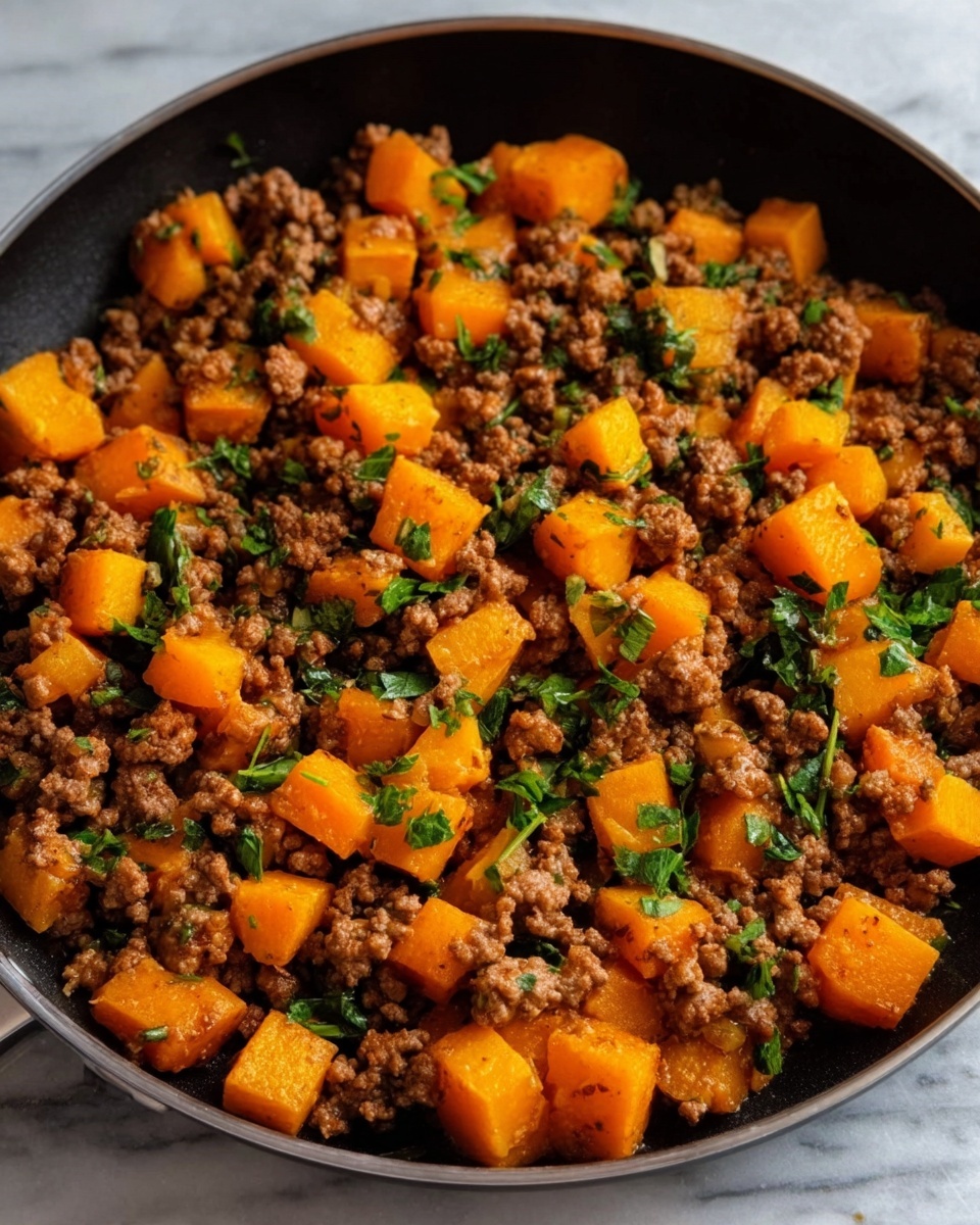 The image shows a close-up of a dark pan filled with cooked ground beef mixed with bright orange cubes of cooked squash. There are small green leafy bits scattered evenly on top of the beef and squash mix. The colors stand out against the pan, with the brown beef and vibrant orange squash pieces contrasting nicely with the green leaves. The pan is sitting on a white marbled surface. Photo taken with an iphone --ar 4:5 --v 7