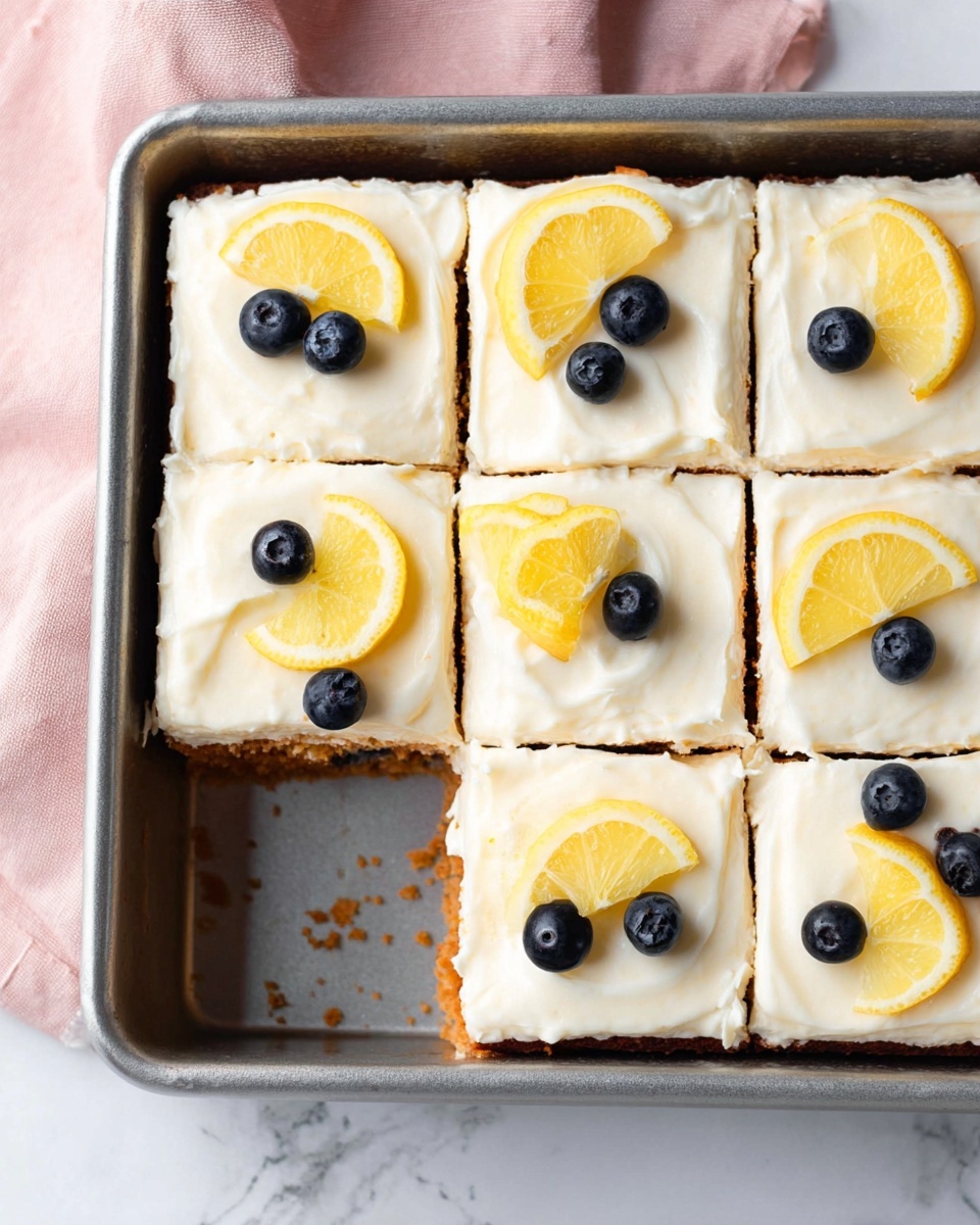 This image shows a metal baking tray with nine square pieces of cake arranged in a 3x3 grid. Each piece has a thick, smooth white cream layer on top, decorated with a small yellow lemon slice and two dark blue blueberries placed near the center of the cream. The cakes are light brown underneath the cream, visible where one piece is missing from the bottom left corner. The tray is on a white marbled surface with a soft pink cloth visible on the left side. The photo taken with an iphone --ar 4:5 --v 7