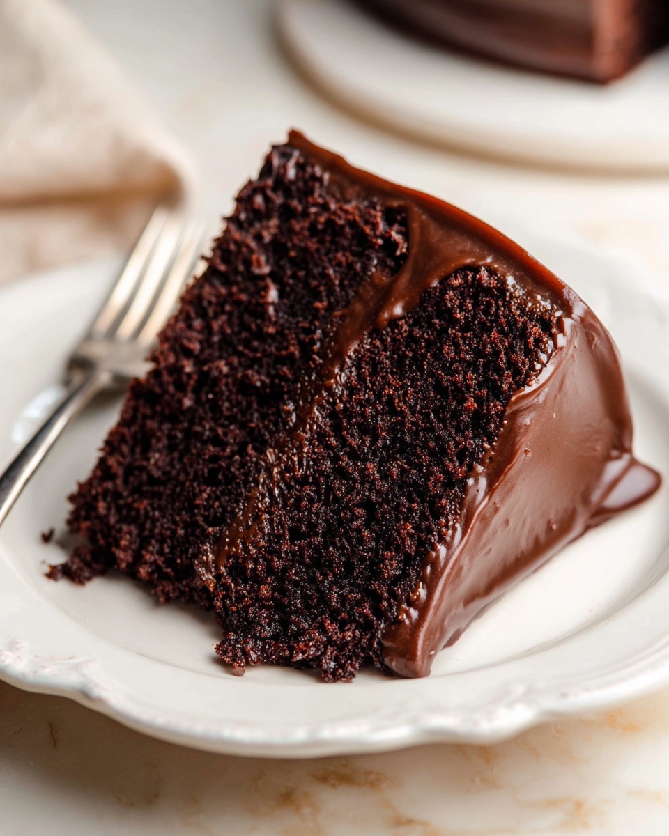 The image shows a slice of two-layer dark chocolate cake on a white plate with a scalloped edge, set on a white marbled surface. The cake layers look moist and dense with a rich, dark brown color, while the outer side and the top are covered with smooth, thick, glossy chocolate frosting that flows slightly over the edges. In the background, a silver fork is partially visible and slightly out of focus, adding to the simple and elegant presentation. Photo taken with an iphone --ar 4:5 --v 7