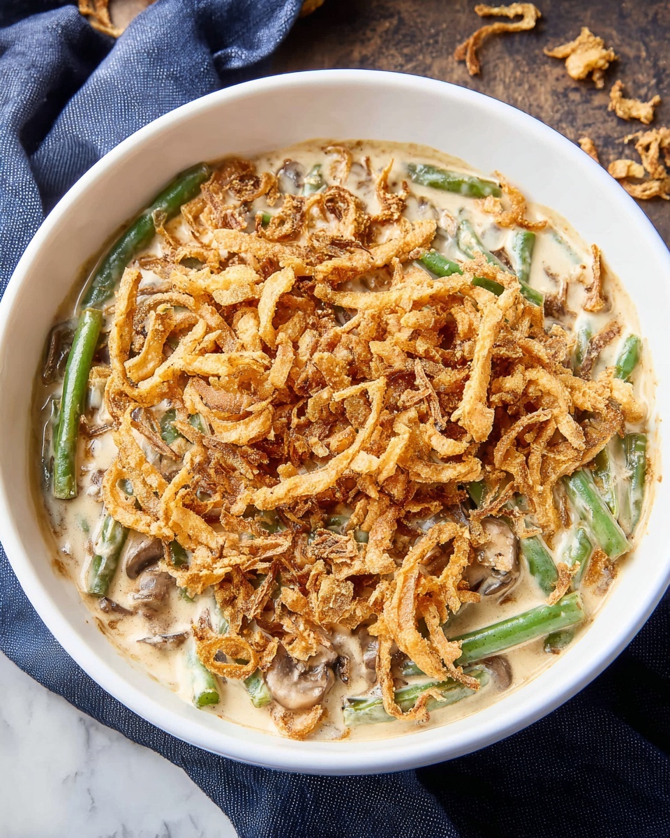 A round white bowl filled with a creamy mixture of light beige sauce with visible pieces of green beans and mushrooms beneath the surface; on top sits a thick layer of golden-brown crispy fried onion strings, arranged to cover the entire surface evenly. The bowl rests on a white marbled surface partially covered by a dark blue cloth, small bits of fried onions scattered on the side. photo taken with an iphone --ar 4:5 --v 7