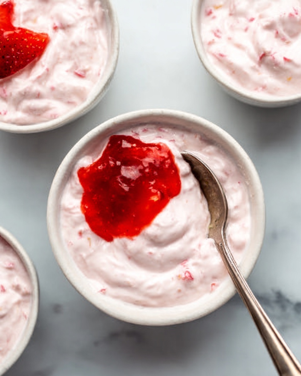 The image shows a close-up of a white bowl filled with pink creamy yogurt mixed evenly with visible small bits of strawberry. On top of the yogurt, there is a bright red strawberry jam layer placed in the left center, with a silver spoon resting on the right side inside the bowl, creating a smooth pattern in the yogurt. In the background, there are two more white bowls with similar yogurt and strawberry jam layers, one positioned at the top left and another slightly blurred in the top right corner, all set on a white marbled surface. The overall color scheme includes soft pinks, bright reds, white, and silver metallic tones. Photo taken with an iphone --ar 4:5 --v 7