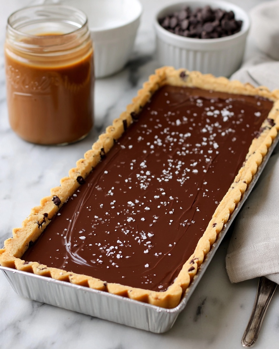 The image shows a long rectangular tart with three layers. The bottom layer is a light brown cookie crust with small dark chocolate chips visible, shaped with a scalloped edge that rises above the tart pan rim. The middle layer is smooth, shiny dark chocolate filling spread evenly inside the cookie crust. The top layer has a few small white salt crystals sprinkled on the chocolate surface. The tart is in a silver metal pan, placed on a white marbled surface. Nearby are a white bowl with small dark chocolate chips and a glass jar with light brown caramel sauce, all softly lit. photo taken with an iphone --ar 4:5 --v 7