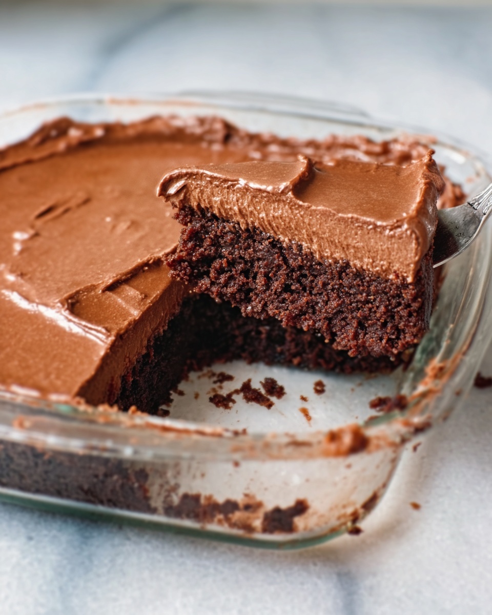 A clear glass dish holds a chocolate dessert with two visible layers; the top layer is smooth and creamy chocolate spread evenly across, with a shiny texture, while the bottom layer is a darker, crumbly chocolate cake base. One slice has been removed, showing the clear division between the layers. There are some small crumbs scattered on the white marbled surface beneath the dish, and a woman's hand is holding a metal spoon with chocolate on it near the left edge of the dish. Photo taken with an iphone --ar 4:5 --v 7