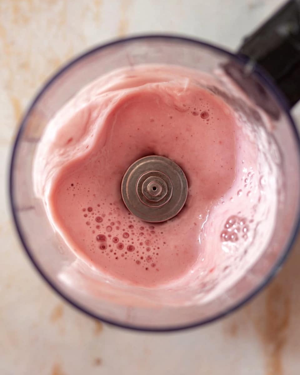 The image shows a close-up view inside a blender jar with a spinning metal blade at the bottom center, blending a light pink creamy mixture. The pink liquid has a smooth, frothy texture with bubbles forming on the surface and around the edges inside the jar. The blender is set on a white marbled surface, and part of a black appliance base is visible on the upper right side. Photo taken with an iphone --ar 4:5 --v 7