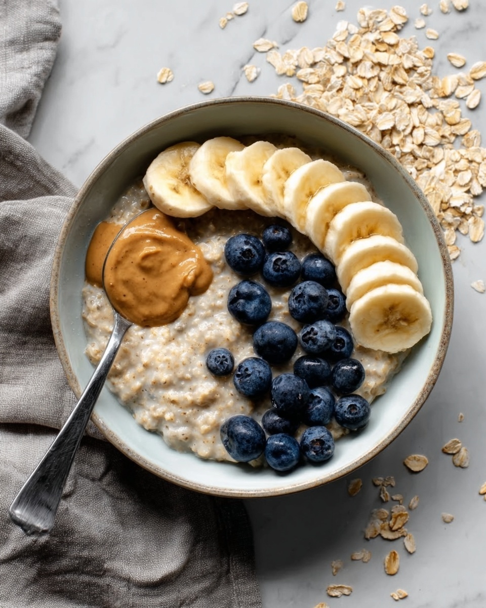 A white bowl filled with creamy oatmeal at the bottom, topped with a neat row of golden banana slices on the upper right side, a small pile of fresh, plump blueberries on the lower right side, and a smooth dollop of light brown peanut butter placed on the left side with a spoon resting inside the bowl. The bowl is set on a white marbled surface with scattered oats and a gray cloth nearby. photo taken with an iphone --ar 4:5 --v 7