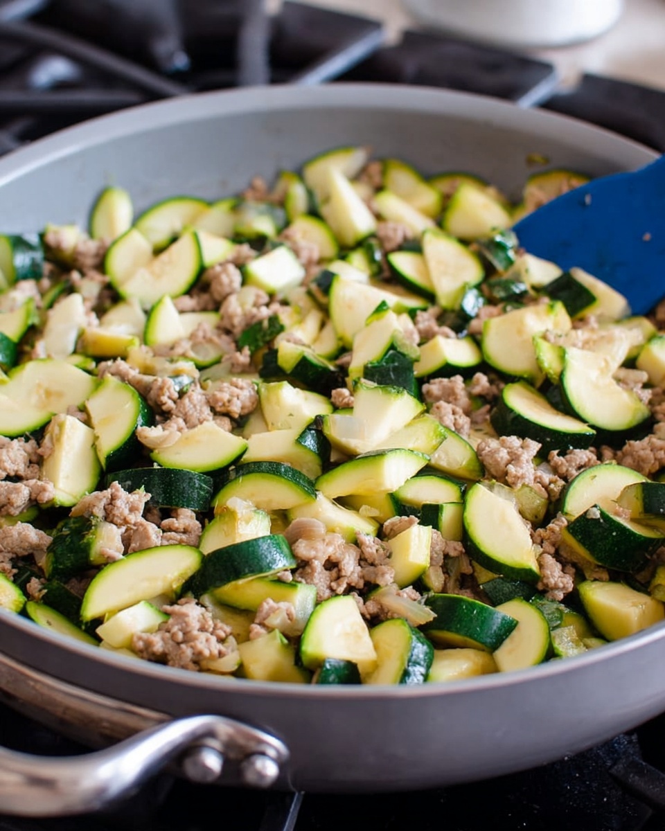 A light grey frying pan filled with a mix of browned, crumbled cooked ground meat and chopped zucchini pieces. The zucchini slices are scattered throughout the pan, showcasing their light green flesh and dark green skin. The meat forms the base layer with small chunks, while zucchini pieces rest on top and throughout, adding contrast with different textures and shades of green. The pan sits on a stove with a silver handle, a blue spatula partially visible on the right side, ready for stirring. Photo taken with an iphone --ar 4:5 --v 7
