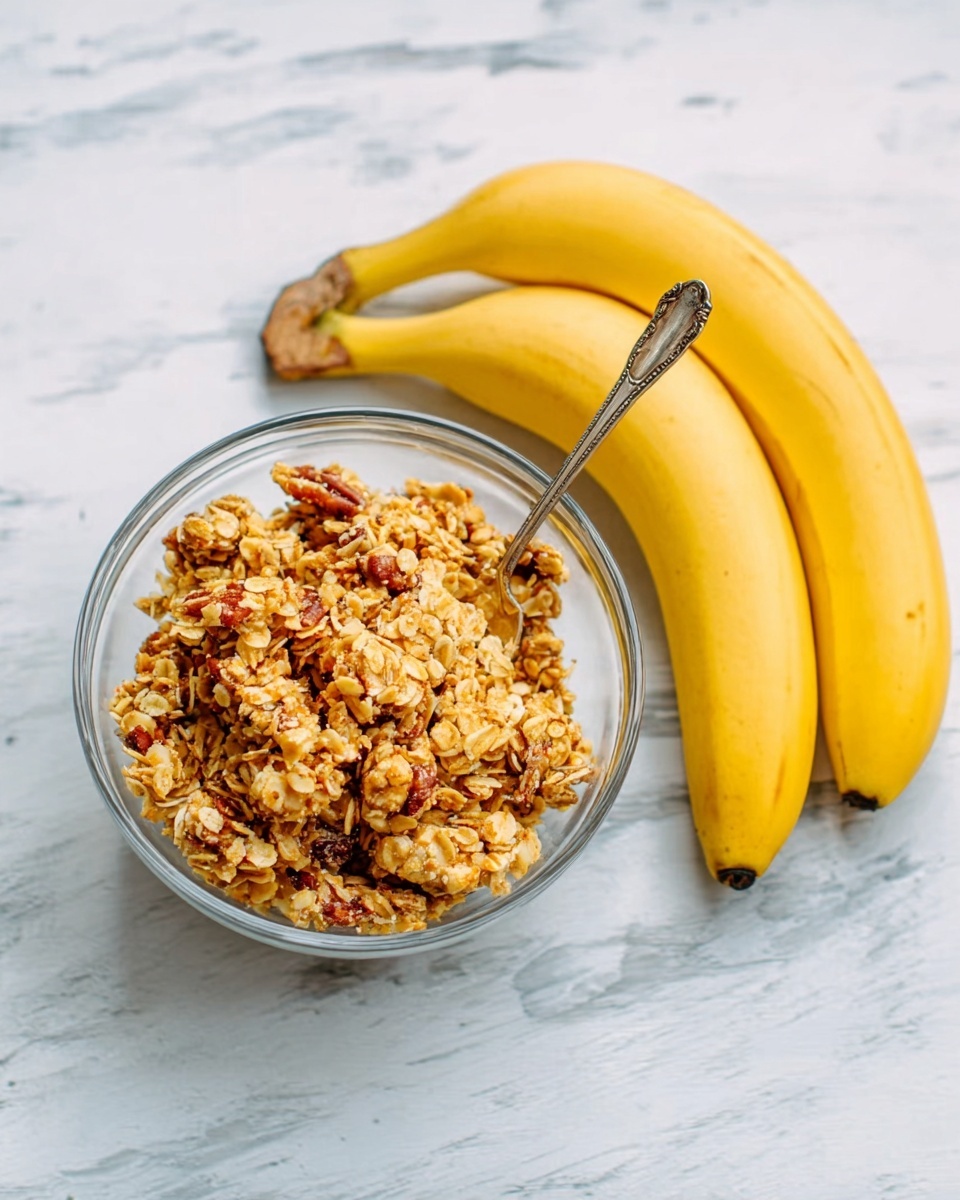 A clear glass bowl filled with chunky granola sits on a white marbled surface. The granola looks crunchy with golden brown clusters, oats, and bits of nuts mixed inside. A silver spoon rests inside the bowl with its handle leaning on the edge. To the right of the bowl, there are three ripe yellow bananas placed closely together with their dark stems facing left. The overall scene is bright with soft natural light. Photo taken with an iphone --ar 4:5 --v 7