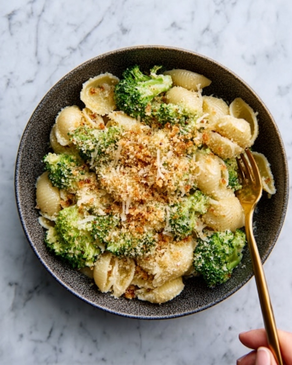 A dark bowl filled with shell pasta mixed with bright green broccoli pieces, topped with a golden-brown breadcrumb sprinkle that adds a rough texture. The pasta is creamy and light yellow, sitting under the crunchy breadcrumbs, and some broccoli sits on top and around the pasta. A gold fork held by a woman's hand rests on the right side of the bowl. The bowl is placed on a white marbled surface. photo taken with an iphone --ar 4:5 --v 7