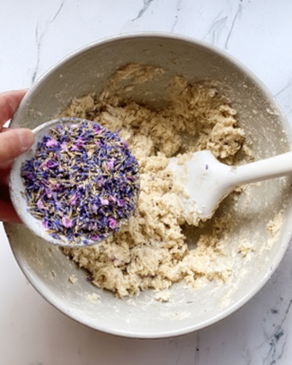 A white bowl filled with small purple flower petals is being held by a woman's hand above a mixing bowl. The mixing bowl contains light beige dough with a rough and sticky texture, and a white spatula is resting inside the dough, partially covered. The background features a white marbled texture. photo taken with an iphone --ar 4:5 --v 7
