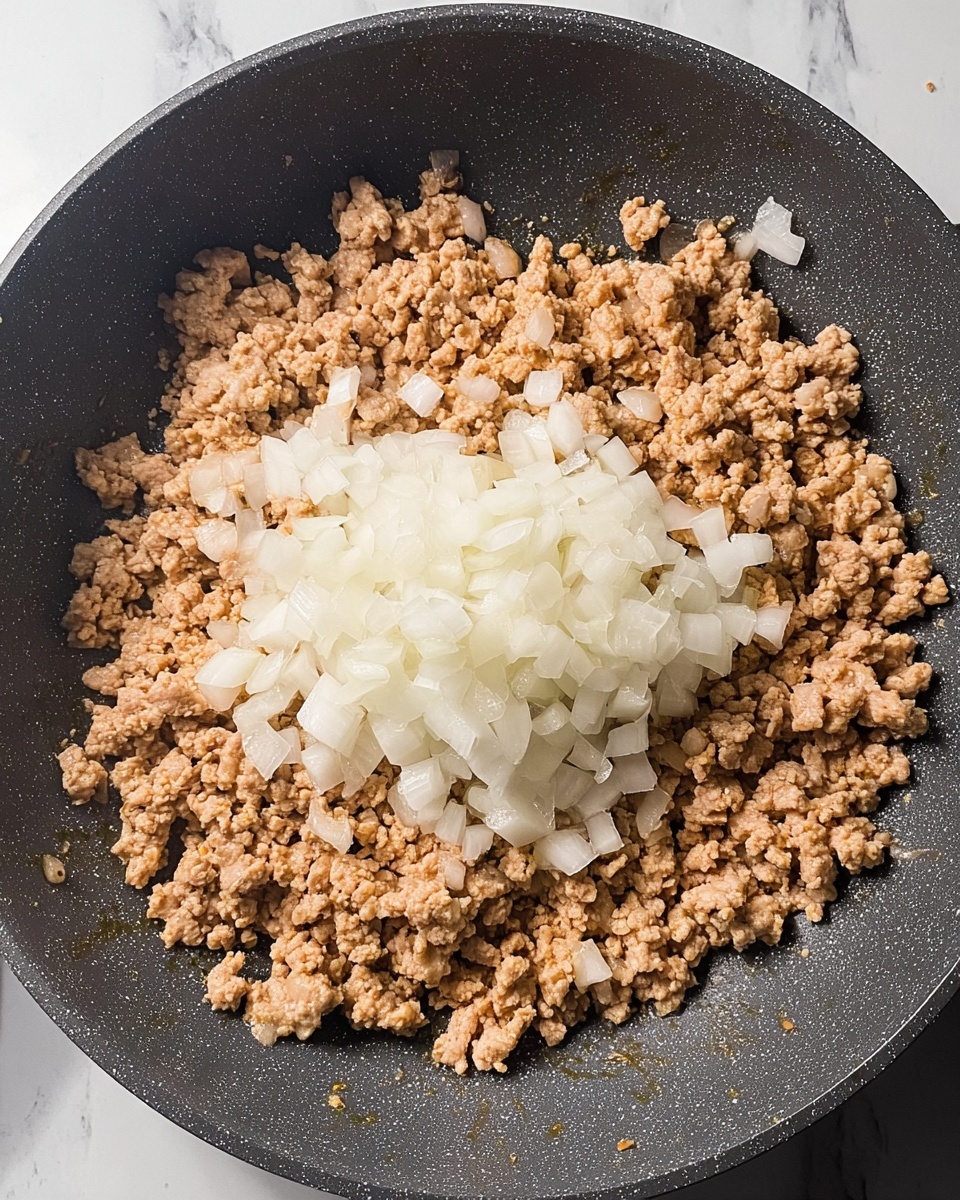 The image shows a frying pan filled with cooked ground meat that is light brown and crumbly in texture, spread evenly across the pan. On top in the center, there is a pile of raw chopped white onions, with their fresh moist look and slightly translucent edges. The pan itself is dark gray with a lightly speckled surface. The background is a white marbled texture. photo taken with an iphone --ar 4:5 --v 7