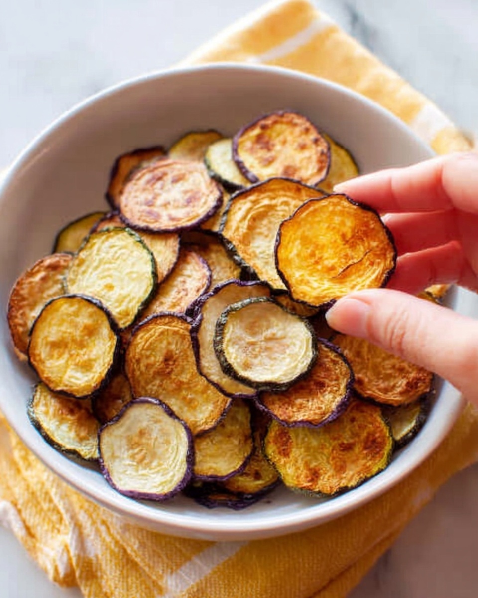 A white bowl filled with many thin, round, crispy slices of cooked eggplant showing different shades of brown and purple, with a woman's hand holding one slice close to the camera, highlighting its golden-brown color and slightly wrinkled texture. The scene is set on a white marbled surface with a yellow striped cloth partially visible under the bowl. Photo taken with an iphone --ar 4:5 --v 7