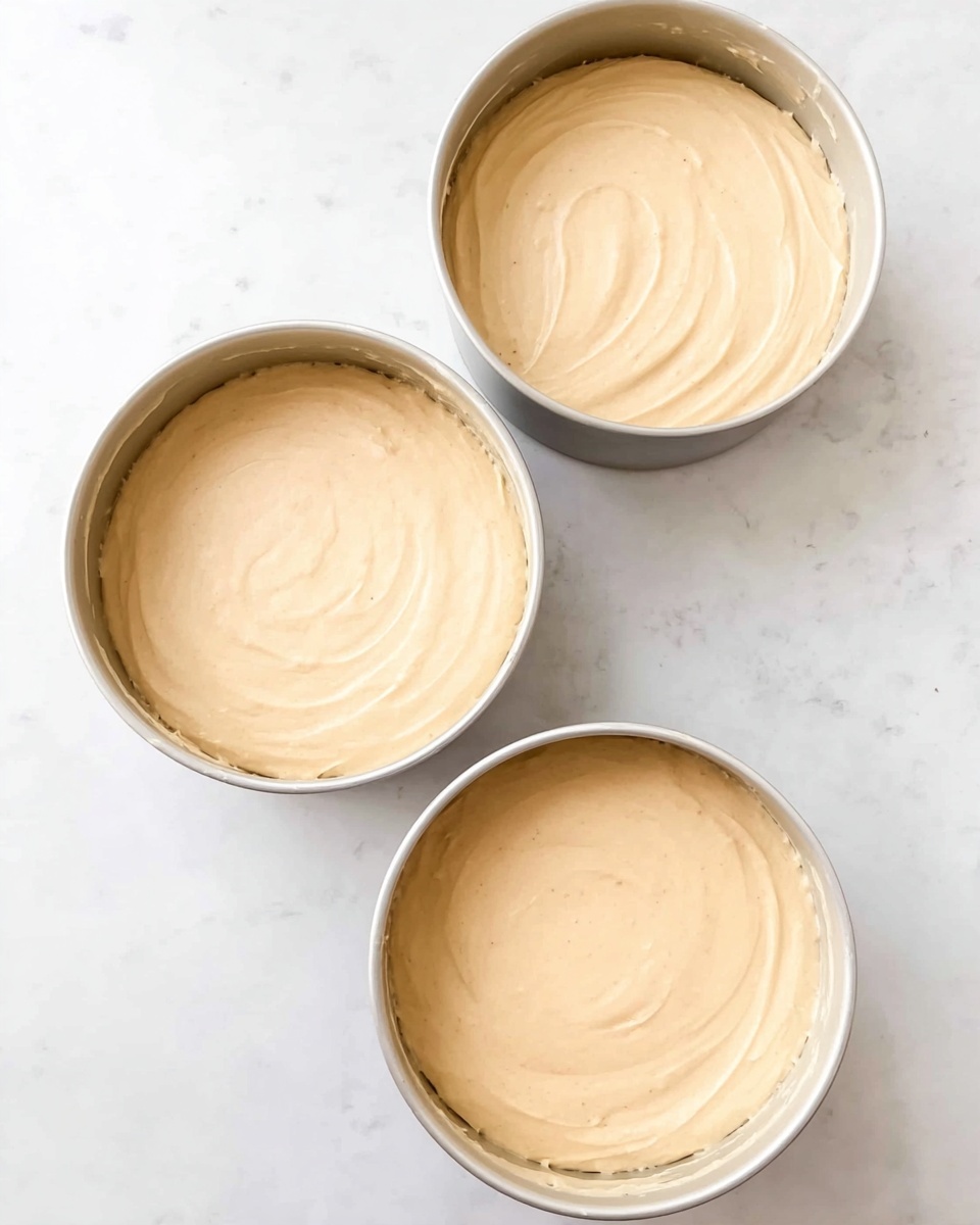 The image shows three round cake pans filled with smooth, light beige batter. Each pan is evenly filled with batter spread flat to the edges, showing a glossy, soft texture on top. The pans are placed on a white marbled surface, arranged in a triangle shape with two on the upper side and one at the bottom. The batter looks thick but easy to spread, with subtle swirls visible across the tops. photo taken with an iphone --ar 4:5 --v 7
