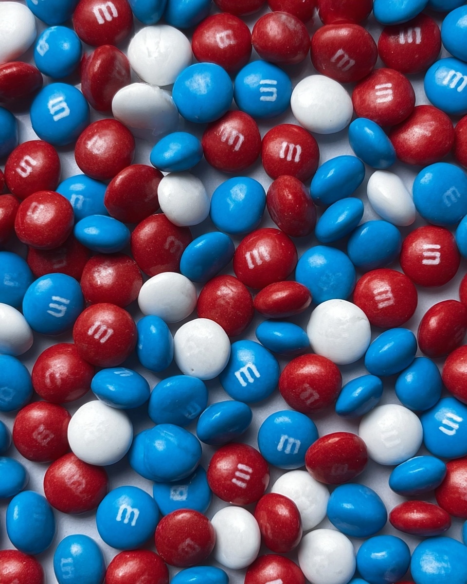 The image shows a close-up pile of small round candies with a smooth, shiny texture. The candies are in three colors: red, blue, and white, mixed evenly throughout the pile. Each candy has a small white letter