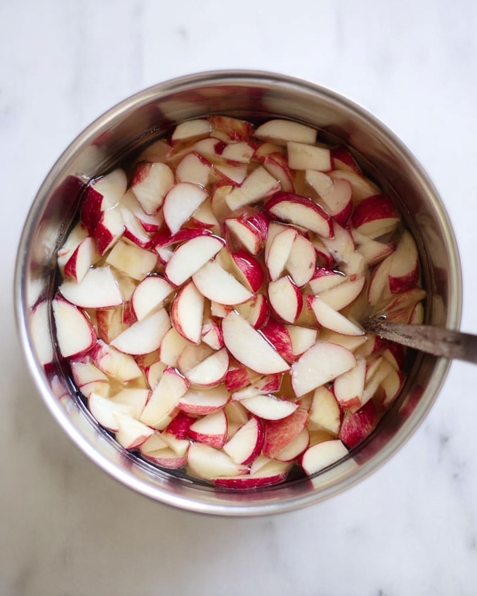 A top view of a silver metal bowl filled with many small pieces of apple with red skin and white flesh, mixed in clear water. The apple pieces are thin and uneven, floating and partially submerged in the water. The bowl sits on a white marbled surface, and a spoon handle is visible resting inside the bowl on the right side. photo taken with an iphone --ar 4:5 --v 7