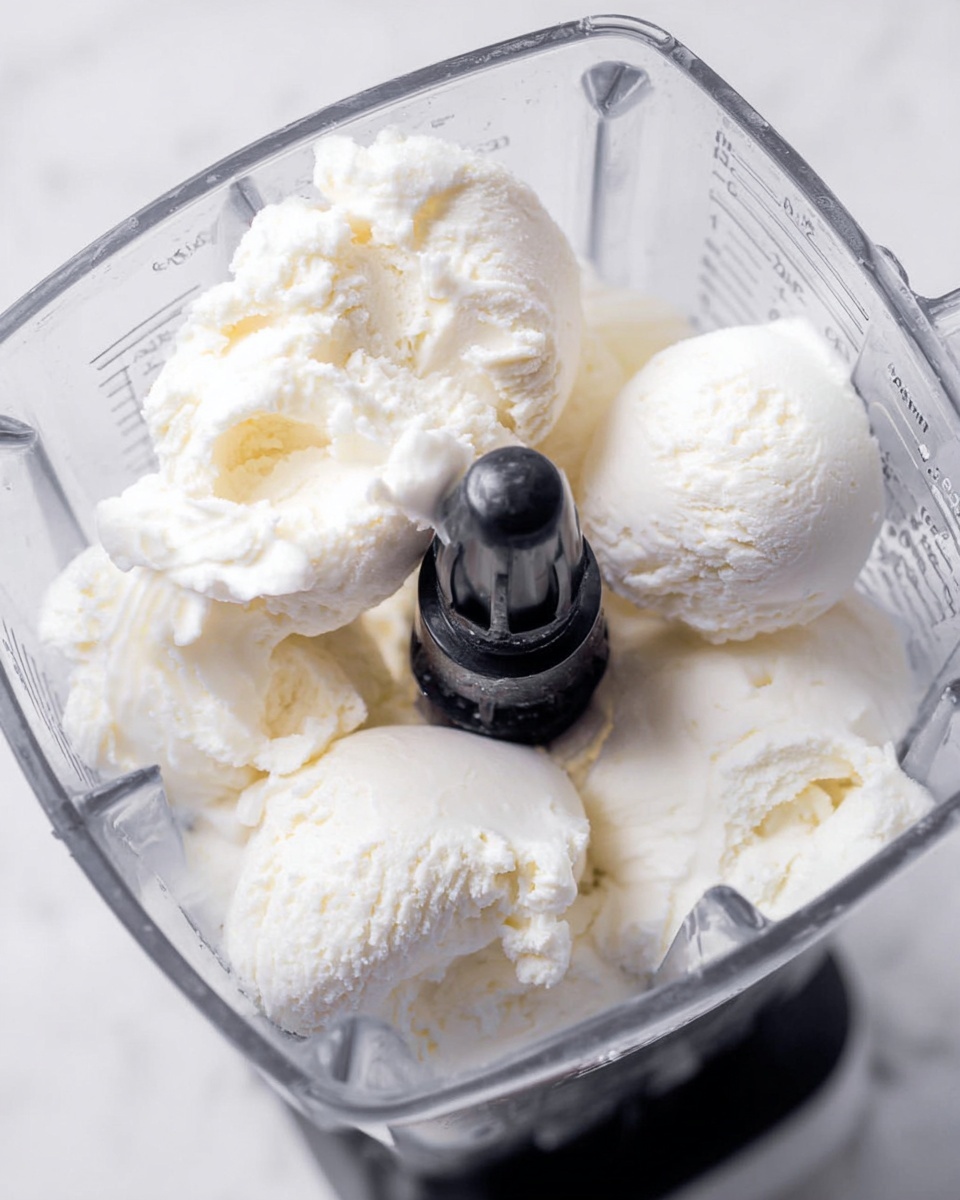 The image shows a clear blender container filled with several large scoops of white ice cream resting around a central black blender blade. The creamy texture of the ice cream is fluffy and soft, with some small air pockets visible. The container has measurement marks on the side and the background is a white marbled texture. Photo taken with an iphone --ar 4:5 --v 7