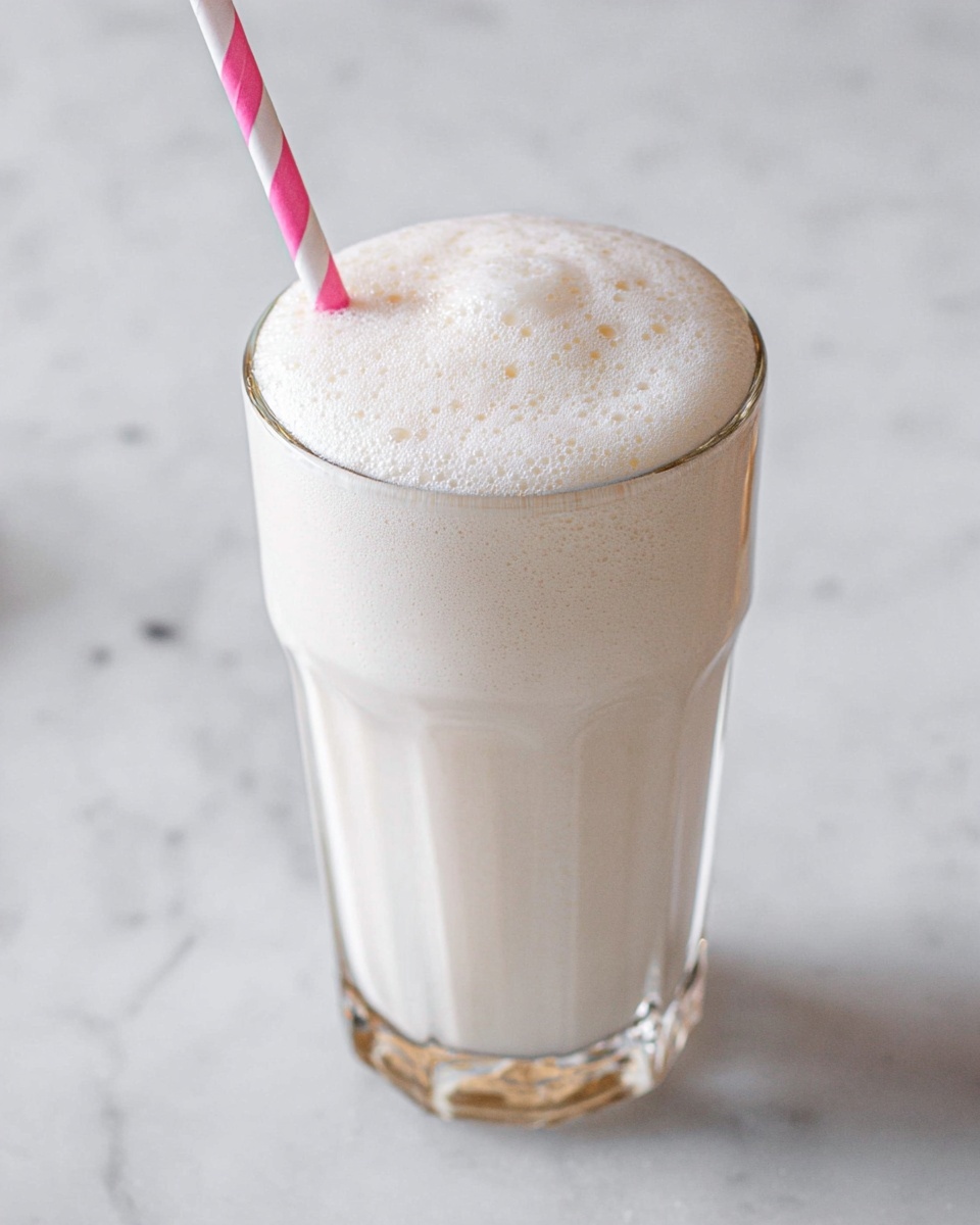 A tall clear glass filled with a creamy white milkshake topped with a thick layer of smooth, frothy foam. The foam has small bubbles around its edges and a slight dome shape at the top. A pink and white striped paper straw is placed on the left side of the glass, reaching down into the milkshake. The glass sits on a white marbled surface. photo taken with an iphone --ar 4:5 --v 7