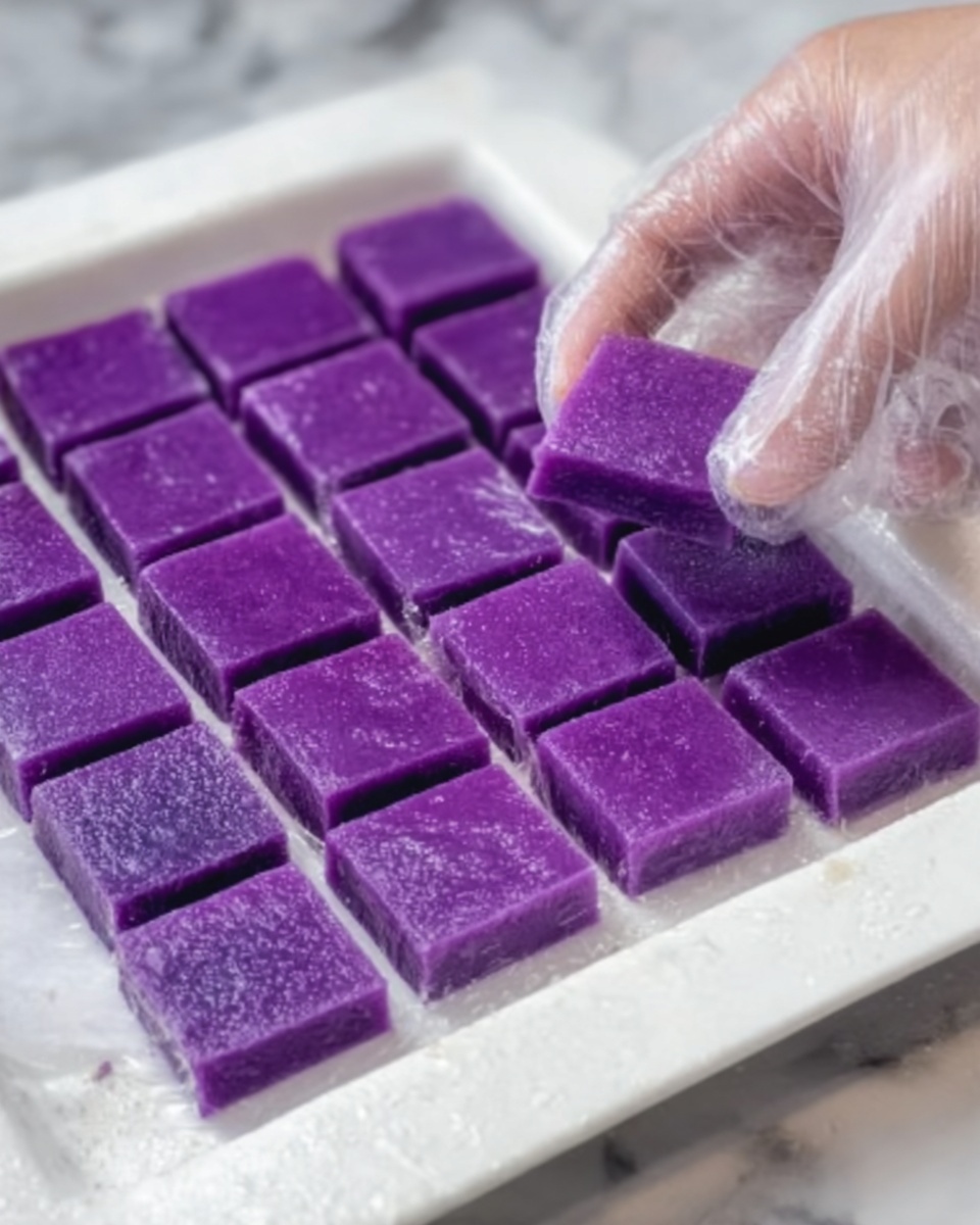 The image shows a white tray with 20 small square purple pieces of a soft dessert neatly arranged in four rows and five columns. Each piece is smooth and shows a dense, slightly sticky texture with a consistent rich purple color throughout. A woman's hand wearing a clear plastic glove is holding one square piece close to the tray, revealing its thickness of about half an inch. The tray is placed on a white marbled surface. Photo taken with an iphone --ar 4:5 --v 7