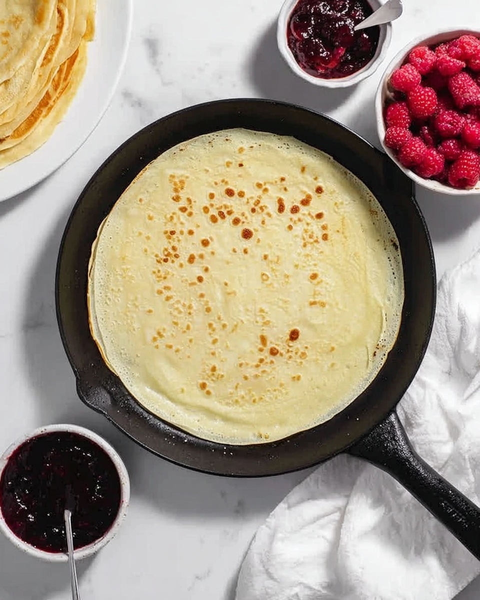 In the image, there is a single light yellow crepe with small brown spots cooking in a black cast iron pan placed on a white marbled surface. To the right of the pan, there are two small white bowls, one filled with bright red fresh raspberries and the other with dark red jam that has a silver spoon inside. At the top left corner of the image, part of another crepe on a white plate is slightly visible. The whole scene is bright and clean with natural light. Photo taken with an iphone --ar 4:5 --v 7
