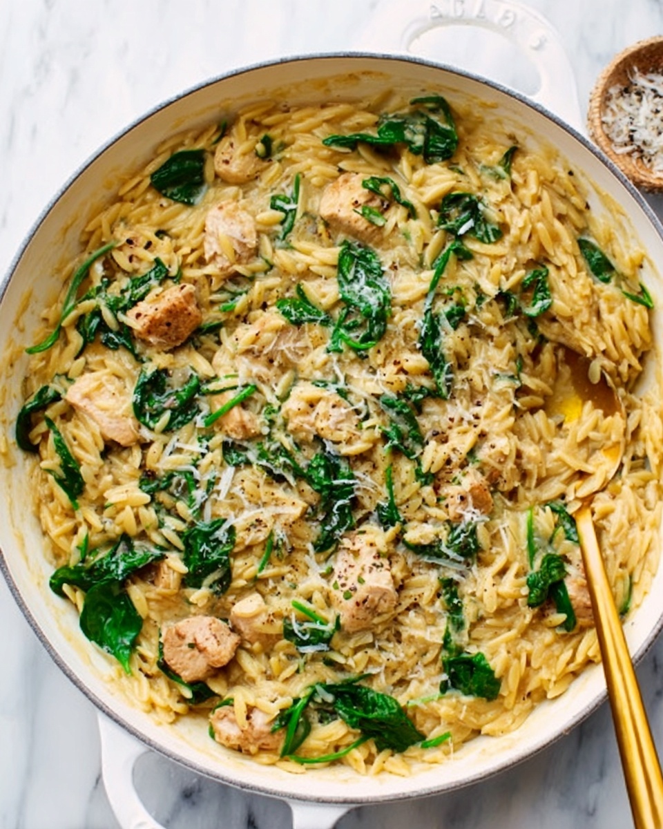 A white shallow pan filled with a creamy orzo pasta dish. The pasta is mixed with pieces of cooked chicken, visible in smaller chunks throughout. Bright green spinach leaves are scattered on top, adding a fresh color contrast. The dish has a slightly saucy texture with some grated cheese sprinkled over it, giving a light, crumbly appearance. A golden spoon is placed inside the pan on the right side. The pan sits on a white marbled surface. Photo taken with an iphone --ar 4:5 --v 7