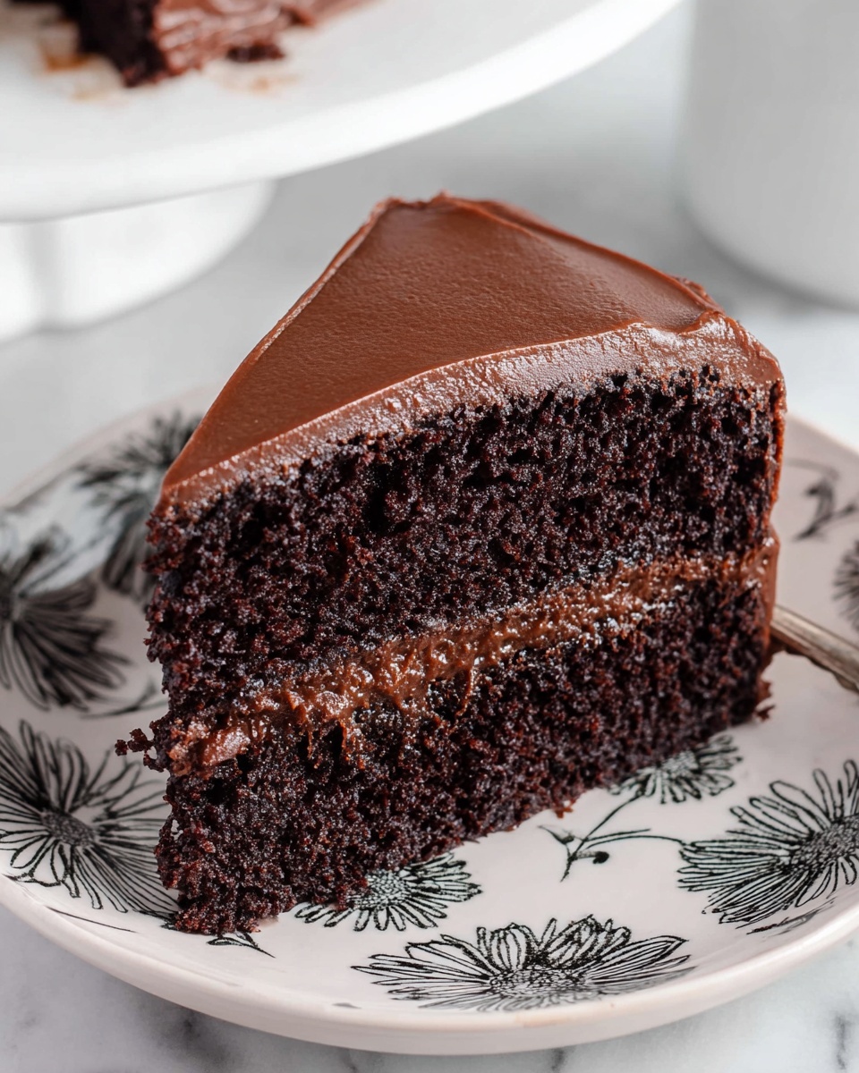 A close-up of a thick two-layer chocolate cake slice with dark, rich chocolate sponge layers separated by a smooth, creamy chocolate frosting layer in the middle. The top and side of the slice are covered in an even, glossy layer of chocolate frosting, showing a slight shine and smooth texture. The slice sits on a white plate with black floral and leaf designs. The background is a white marbled surface with soft lighting highlighting the cake’s moist texture and rich chocolate color. Photo taken with an iphone --ar 4:5 --v 7