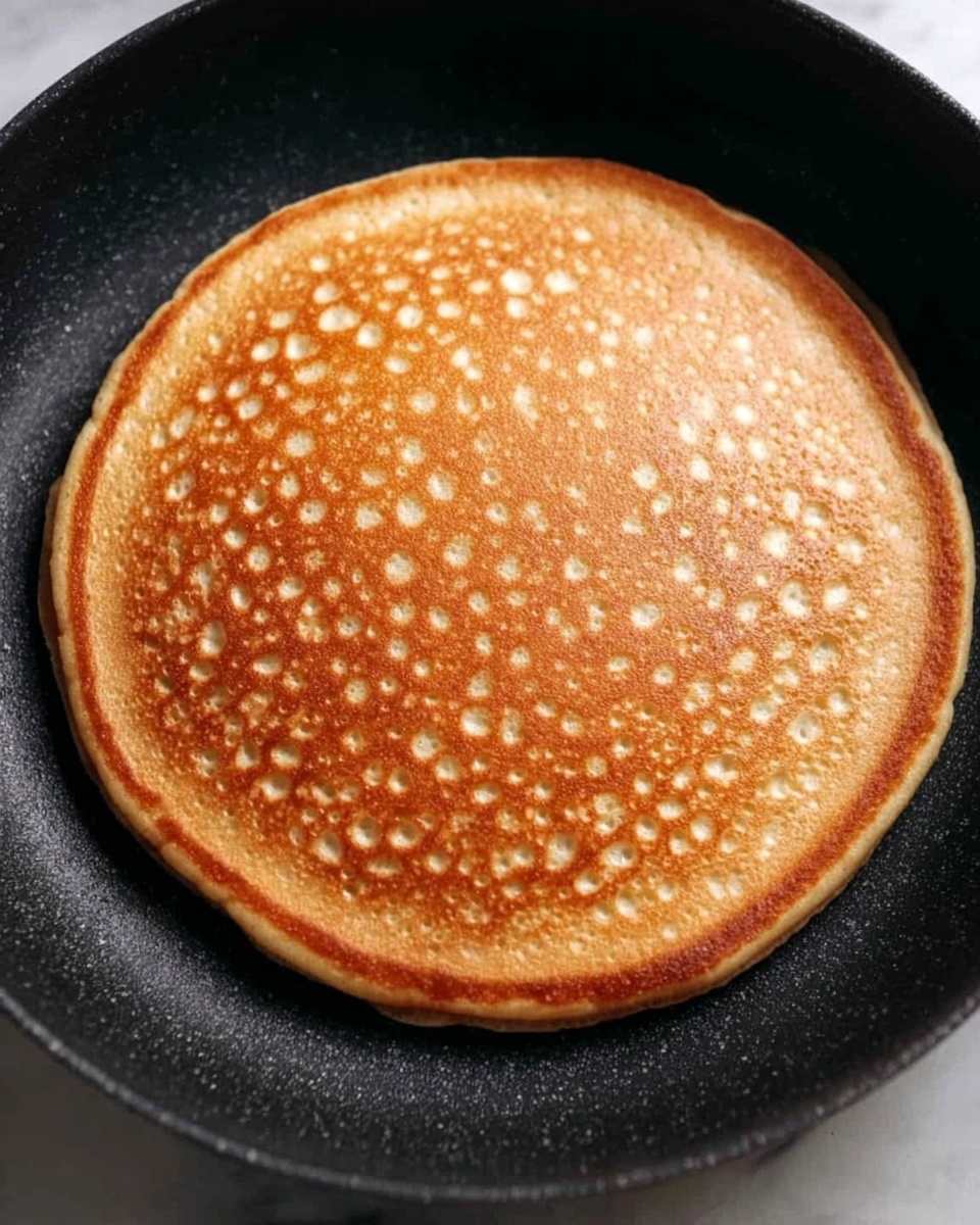 A single golden brown pancake with small round bubbles scattered evenly across the surface is cooking in a black pan. The pancake is thick and has a smooth, slightly porous texture, showing even cooking with a warm caramel color. The pan’s edge is visible around the pancake, and the background is a white marbled texture. photo taken with an iphone --ar 4:5 --v 7
