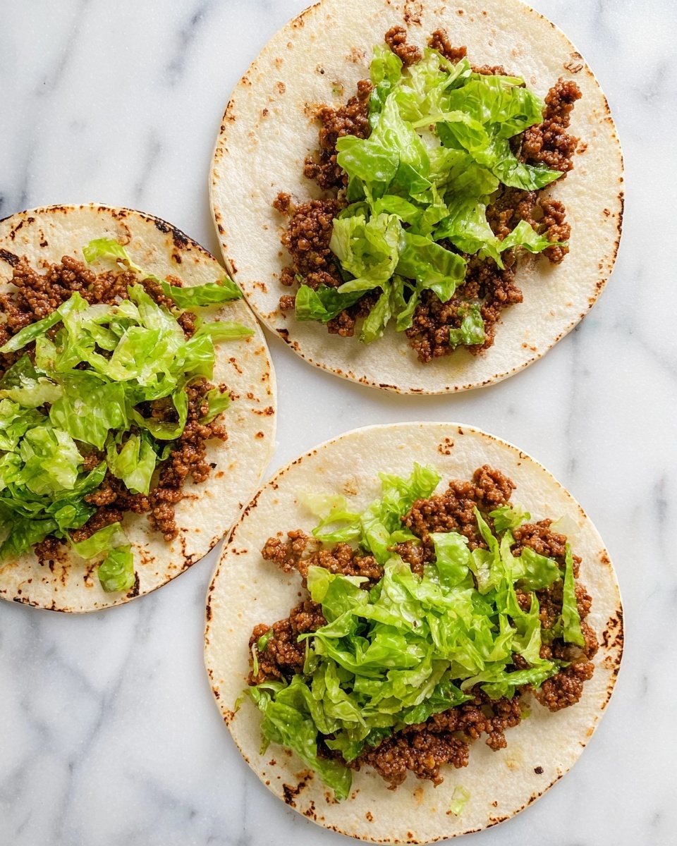 Three small white tortillas lie flat on a white marbled surface. Each tortilla is topped with one layer of crumbly, cooked brown ground meat, evenly spread in the center, and a second layer of fresh, thinly sliced green lettuce scattered over the meat. The tortillas have light brown char marks around the edges. The overall look is simple and fresh with earthy brown and green colors contrasting against the white tortillas and background. Photo taken with an iphone --ar 4:5 --v 7