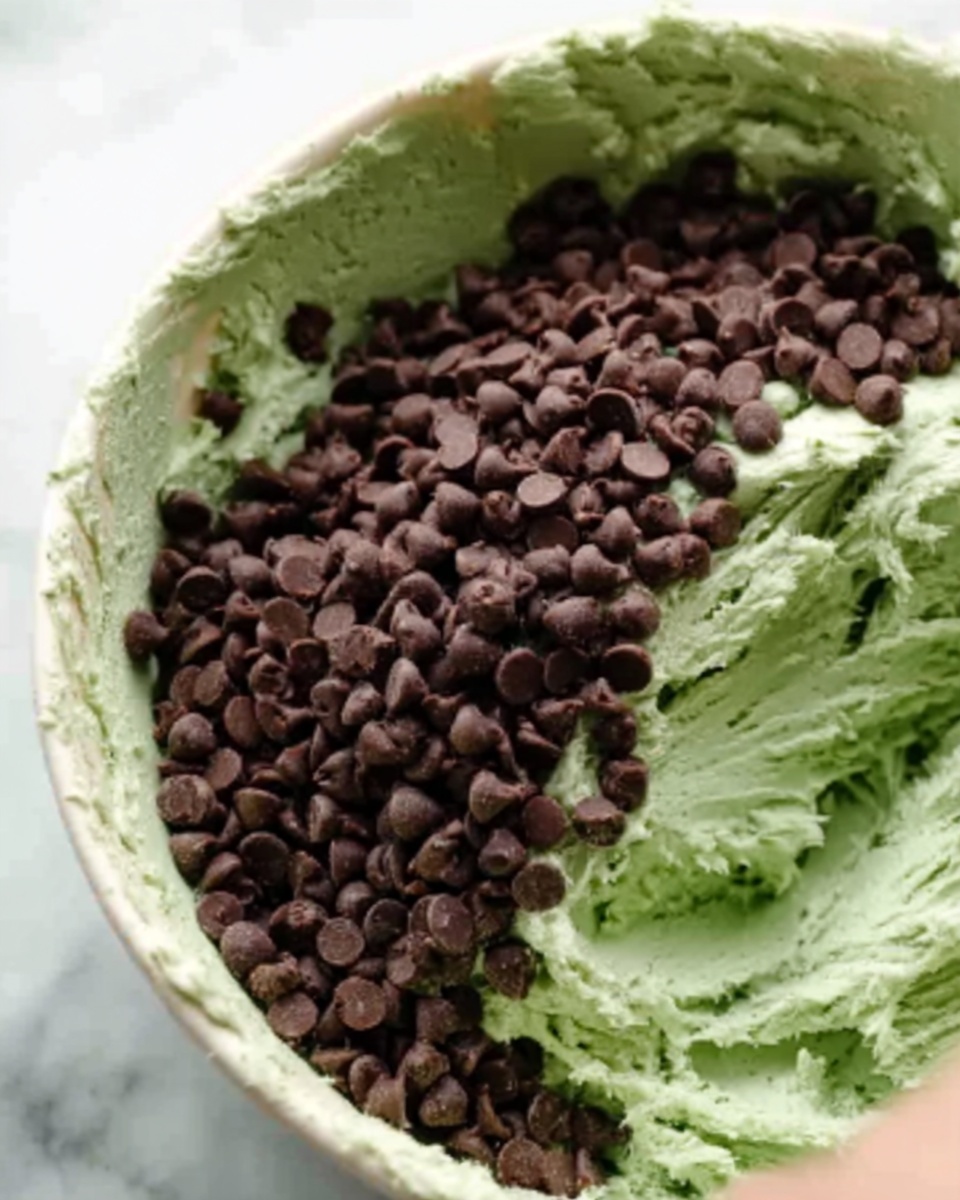 The image shows a close-up of a mixing bowl filled with a thick, pale green dough or batter with a smooth texture. On top of the green mixture, there is a generous layer of dark brown chocolate chips evenly spread over it. Part of a woman's hand is visible near the edge of the bowl, stirring or mixing the ingredients. The bowl sits on a white marbled surface. photo taken with an iphone --ar 4:5 --v 7
