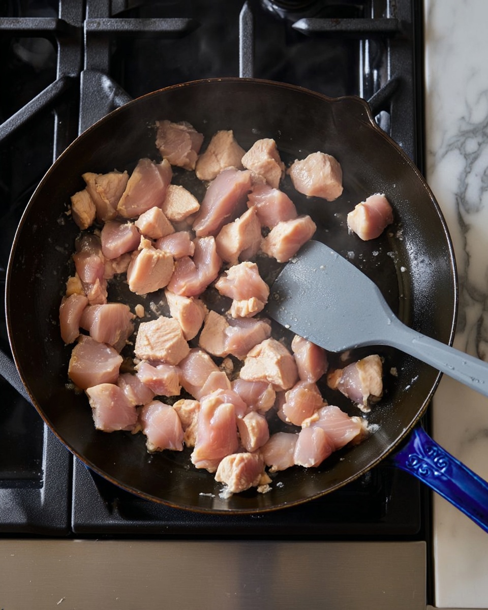 A cast iron pan filled with many small pieces of raw, light pink chicken spread across its surface, some gently steaming as they cook on a black stove. A gray spatula is inside the pan, pushing the chicken pieces slightly. The pan is dark with a shiny blue handle, and the stove has black grates under the pan. To the right of the stove, there is a white marbled countertop. The overall scene shows the beginning stages of cooking chicken chunks. photo taken with an iphone --ar 4:5 --v 7