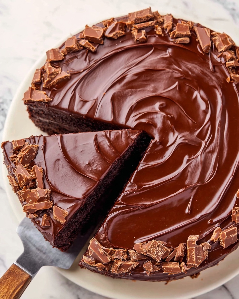 The image shows a round chocolate cake with glossy dark brown chocolate frosting smoothly spread on top in swirled patterns. There is one slice carefully taken out, revealing the cake's thick, rich chocolate layer beneath the frosting. Along the outer edge of the cake is a ring of broken chocolate candy pieces adding texture and extra color contrast. The cake rests on a white plate, sitting on a white marbled surface. A metal spatula is lifting the removed slice. The overall look is rich, smooth, and indulgent. photo taken with an iphone --ar 4:5 --v 7