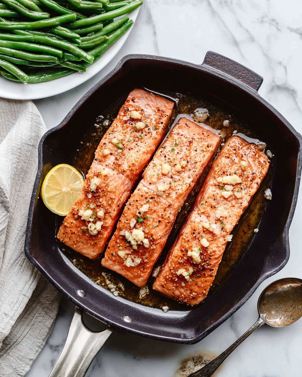 A black pan sits on a white marbled surface, filled with three rectangular salmon fillets placed side by side. The salmon is a light orange-pink color with a slightly shiny and textured surface, topped with small bits of minced garlic and sprinkled with black pepper. A lemon slice rests on the left side inside the pan. To the top left of the pan, there is a white plate filled with cooked green beans that have a fresh green color and a slight shine. Near the bottom right corner of the image, a silver spoon with some sauce on it lies on the marble surface. The overall setting is bright and clean. photo taken with an iphone --ar 4:5 --v 7