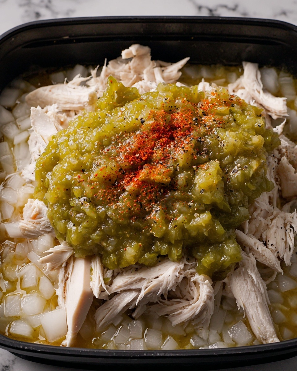 The image shows a close-up of shredded white meat chicken as the bottom layer, with small chopped white onion pieces spread around the base. On top of the chicken, there is a thick, chunky green sauce made from diced green chili or peppers, covering the middle part of the dish. The green sauce is sprinkled with a fine layer of red chili powder and some black pepper scattered on the surface, adding color contrast. The food is placed inside a black container with a white marbled texture visible in the background. photo taken with an iphone --ar 4:5 --v 7