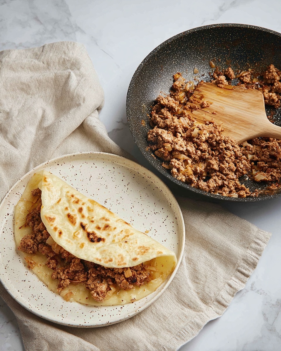 A white speckled plate holds a soft, folded pale yellow flatbread with light brown spots, partially opened to show a layer of crumbly cooked ground meat mixed with small bits of cooked onion inside; next to the plate is a white marbled surface holding a black nonstick frying pan with the same cooked ground meat mixture, a wooden-handled spatula resting in it. A light beige cloth is draped partially over the white marbled surface near the plate. photo taken with an iphone --ar 4:5 --v 7