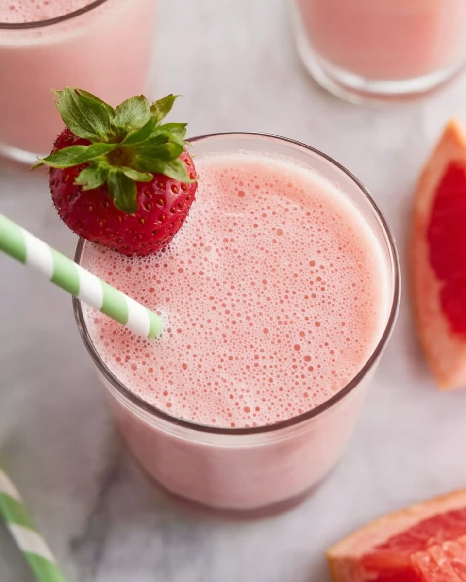 The image shows a clear glass filled with a light pink, frothy smoothie that has a smooth and bubbly texture on top. A fresh red strawberry with green leaves is placed on the rim of the glass on the left side. A green and white striped straw is inserted into the drink near the strawberry. The surface under the glass is a white marbled texture. Part of another glass with the same pink smoothie is visible in the top right corner. A peeled piece of red fruit, likely grapefruit, is partially seen at the bottom right edge. photo taken with an iphone --ar 4:5 --v 7